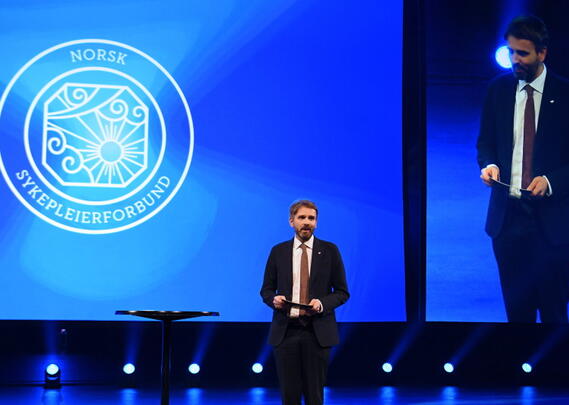 En mann med dress og slips står på en scene med logoen til Norsk Sykepleierforbund bak seg.