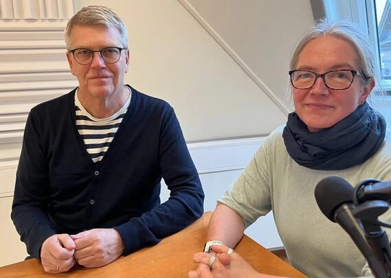 En mann og en kvinne, begge med briller, sitter ved et bord og ser mot kameralinsen.