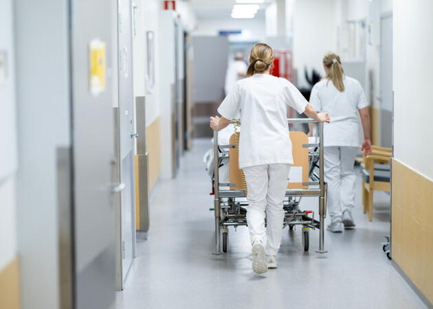 To kvinner i sykepleieruniform ses bakfra trillende på en seng i en sykehuskorridor.