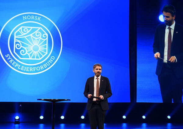 En mann med dress og slips står på en scene med logoen til Norsk Sykepleierforbund bak seg.