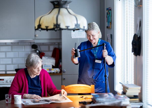 En dame i blå uniform står på kjøkkenet til en eldre dame som sitter på en kjøkkenstol og leser avisen.