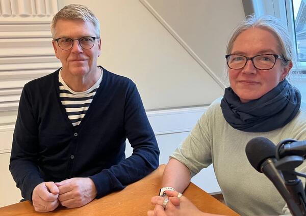 En mann og en kvinne, begge med briller, sitter ved et bord og ser mot kameralinsen.