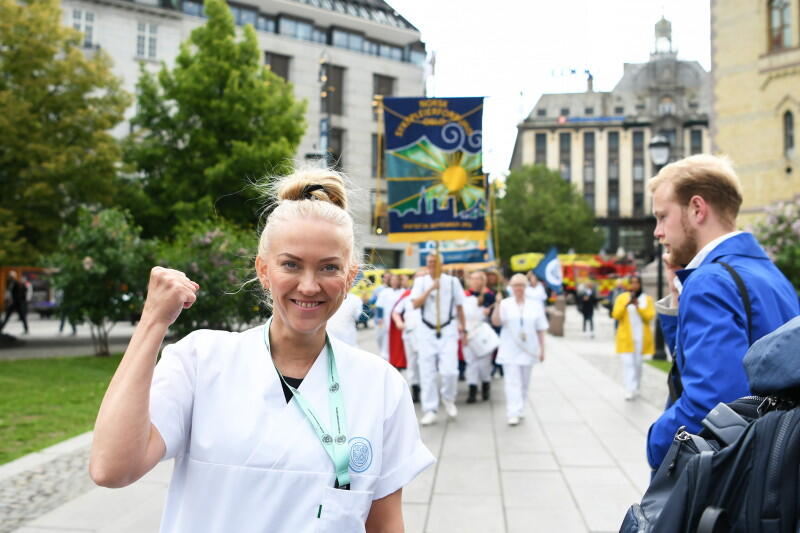 Lill Sverresdatter Larsen i hvit sykepleieruniform smiler til kamera og hever knyttneven. I bakgrunnen ses NSFs fane og sykepleiere i uniform.