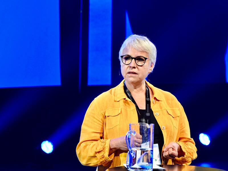 En kvinne med gul jakke, kort hår og svarte briller.