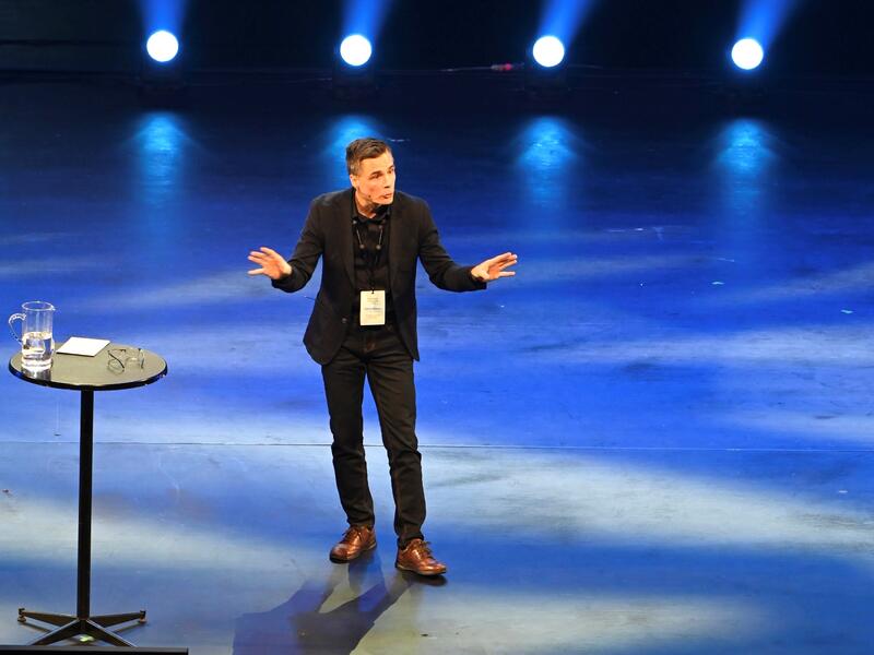 En mann i svart dress gestikulerer på en scene.