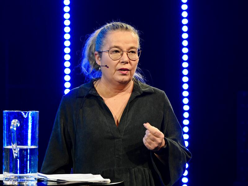 En kvinne med briller, hestehale og mygg foreholder på en scene.