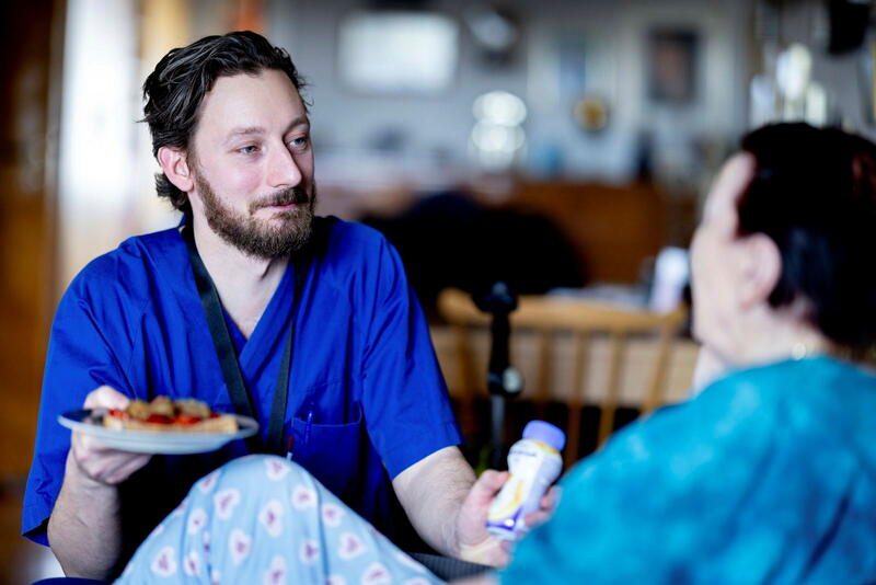 Hjemmesykepleier i blå uniform tilbyr mat til en bruker.