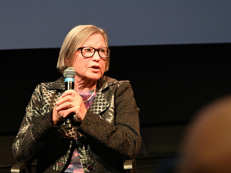 Kvinne med briller snakker i en mikron og ser mot høyre.