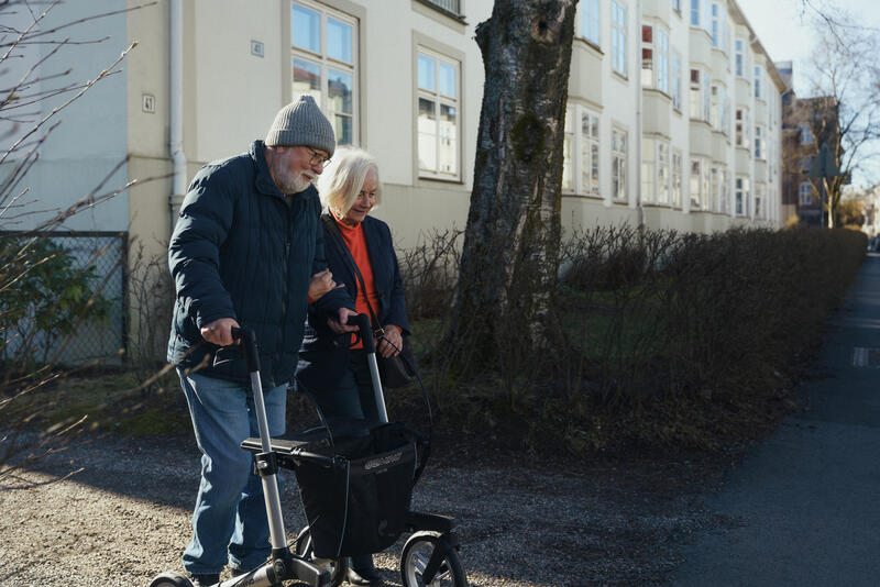 To eldre personer som går arm i arm. Den ene går med rullator. 